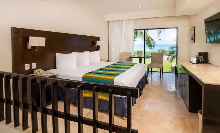 Habitación de hotel con cama grande, área de café, mesa con sillas y balcón con vista tropical al mar.