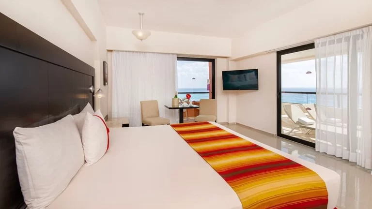 Habitación de hotel con cama grande, mesa con sillas y balcón con vista al mar y paracaidistas en el cielo.