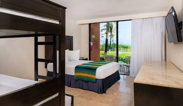 Habitación de hotel con cama queen, litera, televisor y patio con vista al jardín y al mar.